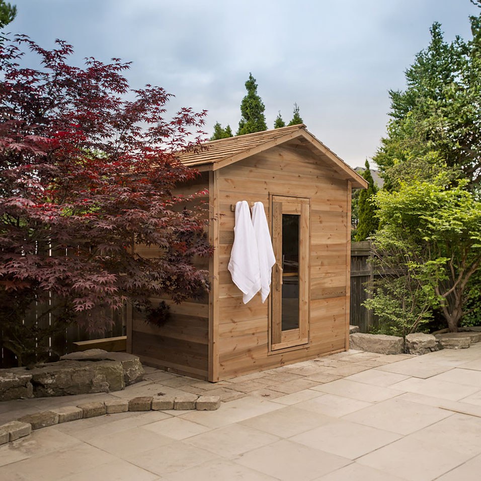 Outdoor Cabin Sauna in knotty red cedar with a glass door and cedar shingle roof on a stone patio.